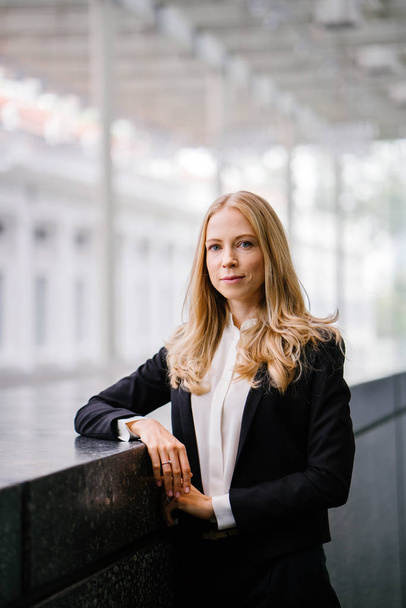 Russian woman in a suit leans against a stone in the day and is smiling. She is blond with blue eyes and is attractive and professional in a black suit and white shirt. - Photo, image