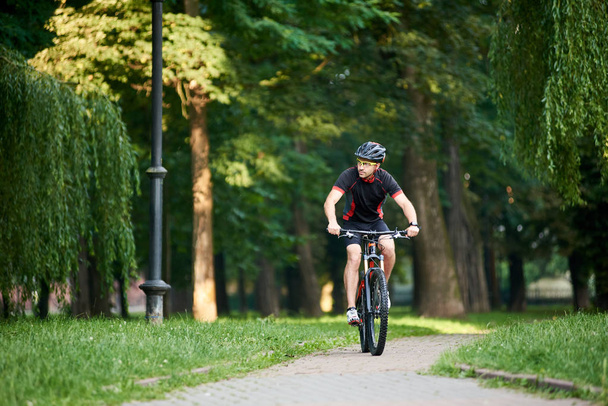 Toned male biker in professional sportswear and protective helmet looking to side while riding bike down park alley on summer day. Exercising, reaching goal - Photo, Image