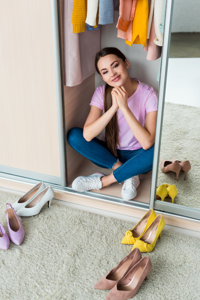attractive young woman sitting inside of cabinet with various shoes on floor at home - Photo, Image