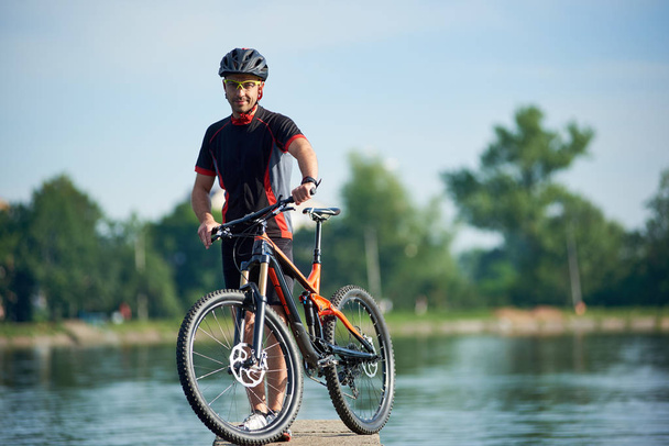 Guapo chico ciclista en ropa de ciclismo y casco posando cerca de la bicicleta mirando a la cámara, sonriendo. Deportista feliz tomando un descanso en el muelle cerca del lago
. - Foto, Imagen