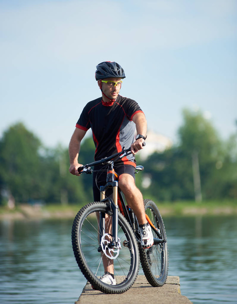 Radler in professioneller Radsportbekleidung und Schutzhelm posieren in der Nähe von Fahrrädern und schauen weg, wenn sie an zukünftige Erfolge und Errungenschaften denken. Sportler macht Pause an Seebrücke am See. - Foto, Bild