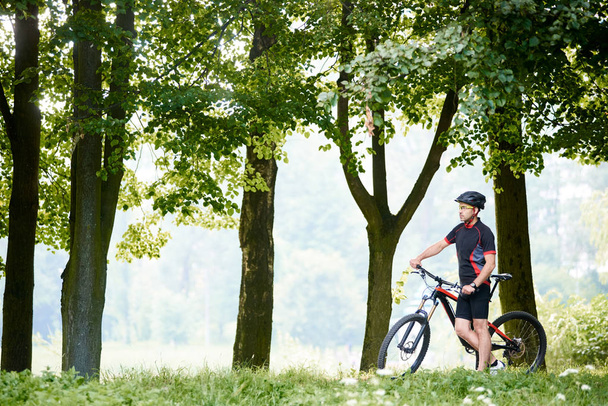 Adam profesyonel bisikletçi bisiklet giyim ve koruyucu kask ayakta Bisiklet yüksek ağaçları arasında güzel güneşli yeşil Park yakınındaki. Sporcu rahatlatıcı. Sağlıklı yaşam, açık havada eğitim kavramı - Fotoğraf, Görsel