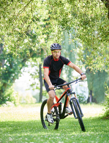 Young guy cyclist in cycling clothes and protective helmet, riding bicycle along green park alleys. Sportsman training outdoors. Concept of healthy lifestyle, connecting with nature - Photo, Image