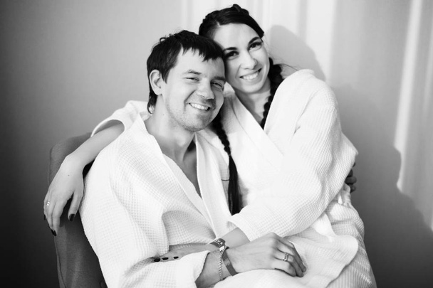 Молодой муж и жена белых халатов играют и улыбаются на кресле в гостиничном номере. Закрывай. Черное и белое
. - Фото, изображение