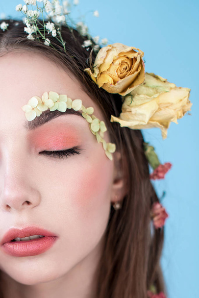 Cosmetics and manicure. Close-up portrait of attractive woman with dry flowers on her face and hair, pastel color, perfect make-up and skin on blue background. Fresh, trendy, spring retouched portrait - Photo, Image