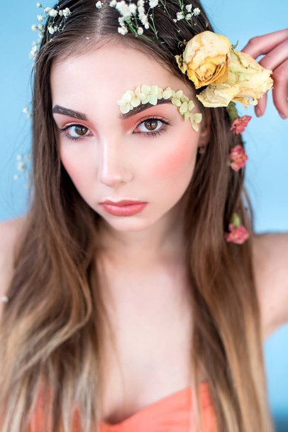 Cosmetics and manicure. Close-up portrait of attractive woman with dry flowers on her face and hair, pastel color, perfect make-up and skin on blue background. Fresh, trendy, spring retouched portrait - Photo, Image