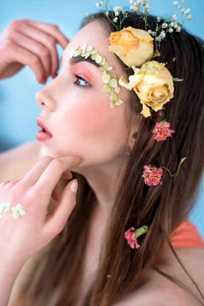 Cosméticos y manicura. Retrato de cerca de una mujer atractiva con flores secas en la cara y el cabello, color pastel, maquillaje perfecto y piel sobre fondo azul. Retrato retocado de primavera fresco, moderno
 - Foto, Imagen