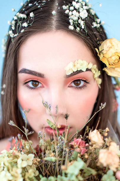 Cosmetica en manicure. Close-up portret van aantrekkelijke vrouw met droge bloemen op haar gezicht en haar, pastel kleur, perfecte make-up en huid op blauwe achtergrond. Frisse, trendy, voorjaar geretoucheerde portret - Foto, afbeelding