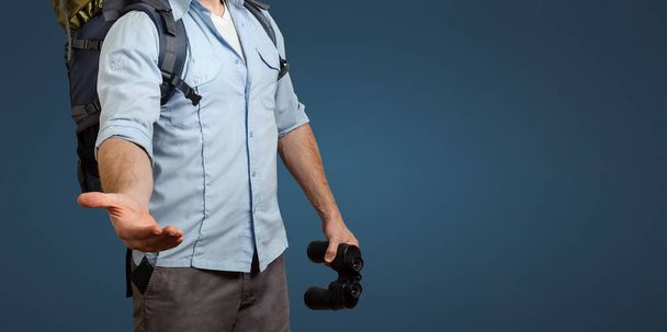 Unrecognizable Young Traveler Man With A Backpack And Binoculars On Blue Background. Stretches Out His Hand. Help In Travel Concept - Photo, image