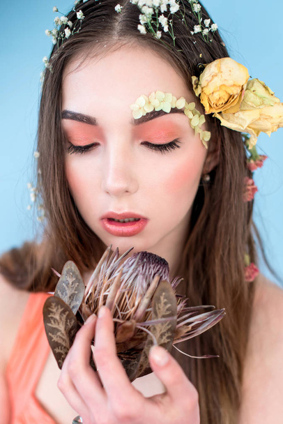 Cosméticos y manicura. Retrato de cerca de una mujer atractiva con flores secas en la cara y el cabello, color pastel, maquillaje perfecto y piel sobre fondo azul. Retrato retocado de primavera fresco, moderno
 - Foto, Imagen