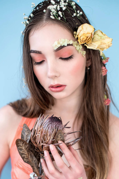 Cosmetics and manicure. Close-up portrait of attractive woman with dry flowers on her face and hair, pastel color, perfect make-up and skin on blue background. Fresh, trendy, spring retouched portrait - 写真・画像