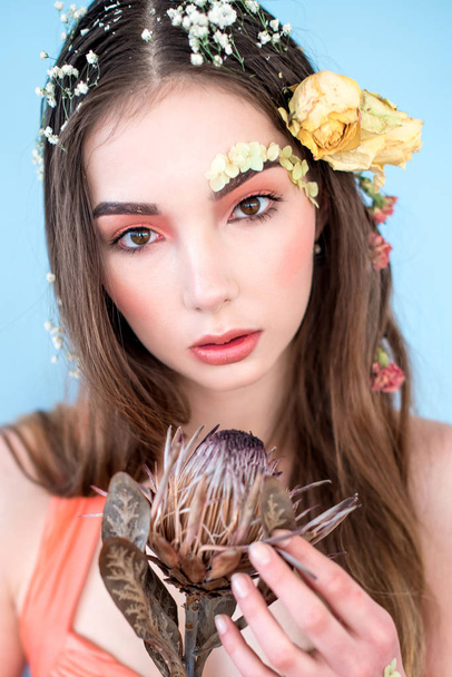 Cosméticos e manicure. Retrato close-up de mulher atraente com flores secas em seu rosto e cabelo, cor pastel, maquiagem perfeita e pele no fundo azul. Retrato retocado de primavera fresco, na moda
 - Foto, Imagem