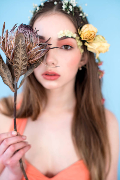 Cosmetics and manicure. Close-up portrait of attractive woman with dry flowers on her face and hair, pastel color, perfect make-up and skin on blue background. Fresh, trendy, spring retouched portrait - 写真・画像