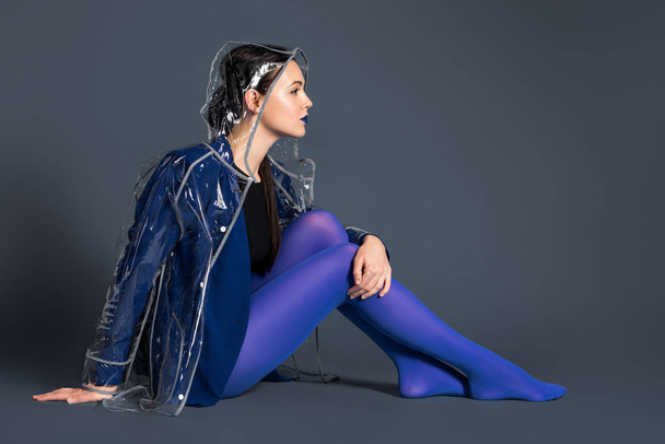 Stylish woman in blue tights and transparent raincoat sitting on dark background - Valokuva, kuva