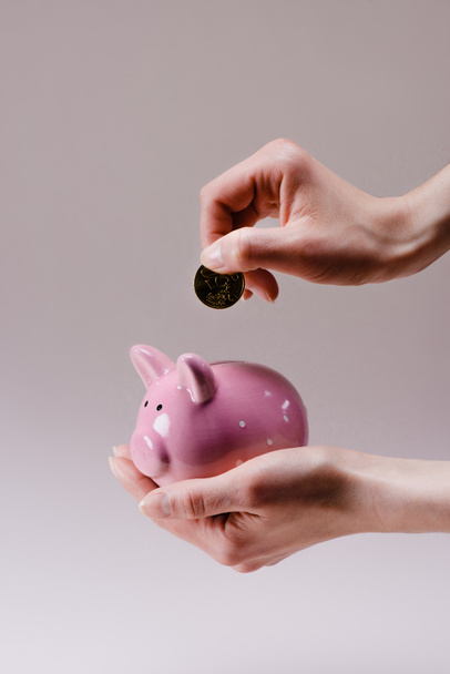 cropped shot of woman putting 50 euro cent into pink piggy bank in hand isolated on lilac - 写真・画像