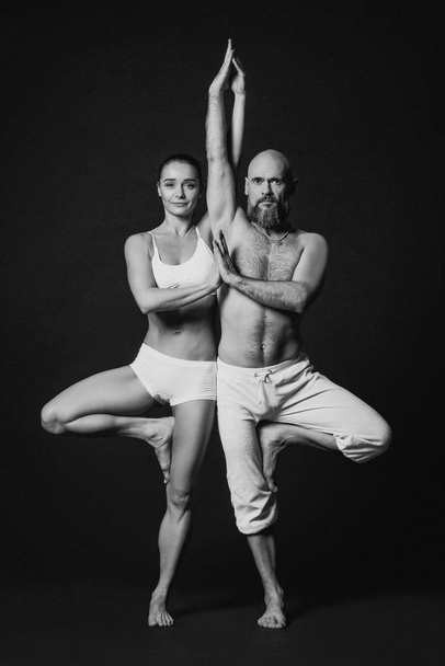 Monochrome portrait of beautiful sporty couple: woman and man in white clothes doing yoga asanas together indoor  - Photo, Image