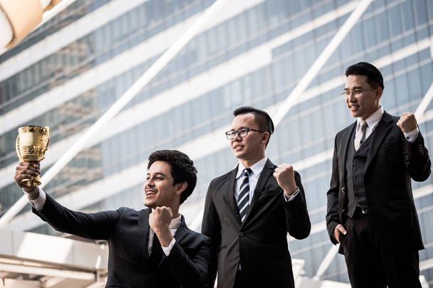 Счастливые азиатские бизнесмены с золотым кубком за победу в бизнес-проекте. Командная работа и концепция успеха
 - Фото, изображение