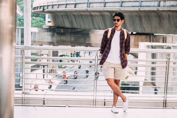 Portrait de jeune homme asiatique en vêtements décontractés avec lunettes de soleil et sac à dos sur fond de scène urbaine
. - Photo, image