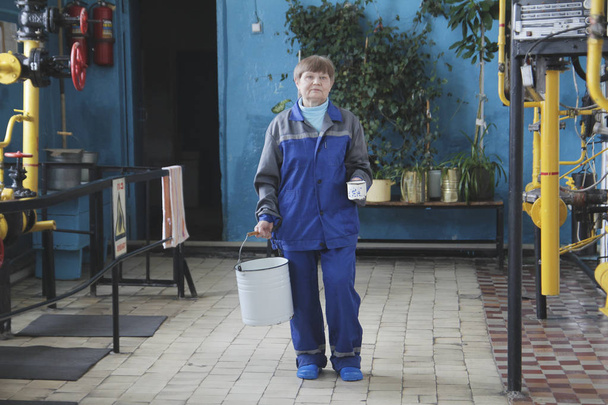 Smiling old woman cleaner with empty bucket in workwear on manufacturing site - 写真・画像