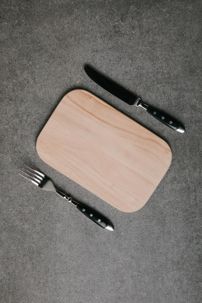 top view of wooden board with fork and knife on grey - Foto, Imagem