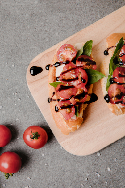 top view of delicious bruschetta with balsamic on wooden board and fresh tomatoes on grey   - Fotó, kép