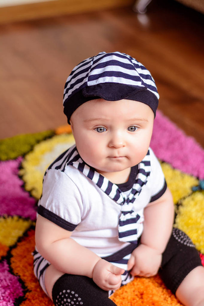 Kaunis vauva raidallinen paita ja hatut istuu matolla huoneessa
 - Valokuva, kuva