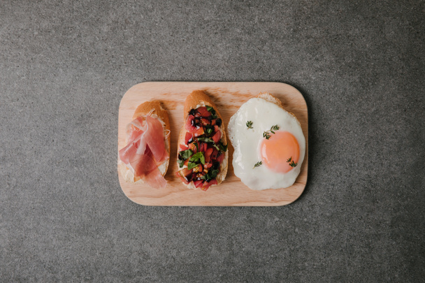 top view of delicious antipasto bruschetta with fried egg on wooden board on grey - Фото, изображение