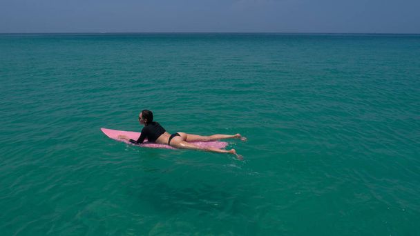 Vista aérea del dron de la hermosa mujer joven en traje de baño y gafas de sol con tabla de surf rosa disfrutando de sus vacaciones en agua de mar clara increíble
 - Foto, imagen