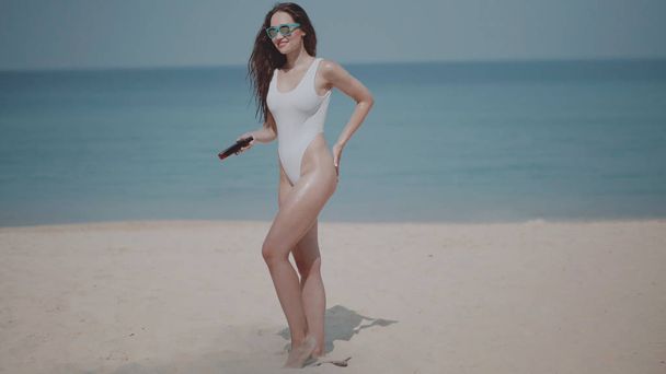 Hermosa chica en traje de baño aplicando crema solar aceite bronceado en el cuerpo en la playa de arena tropical durante las vacaciones de verano sobre el mar y el fondo del cielo
 - Foto, imagen