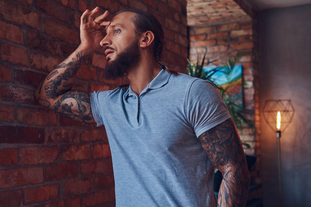 Handsome tattooed male with a stylish haircut and beard, in a gray t-shirt, standing leaning against a brick wall in a room with a loft interior. - Valokuva, kuva