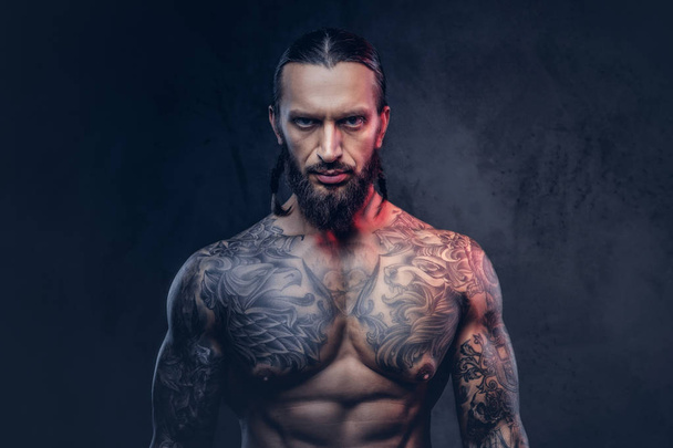 Close-up portrait of a muscular bearded tattooed male with a stylish haircut. Isolated on a dark background. - Photo, Image
