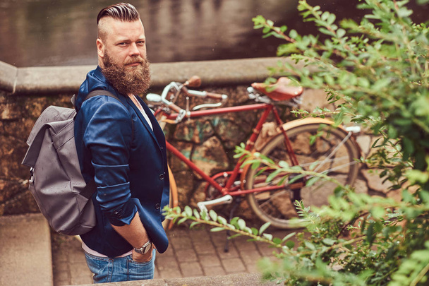 Portrait of a bearded male with a haircut dressed in casual clothes with a backpack, standing in a city park. - 写真・画像