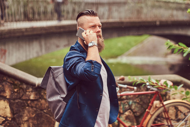 Portrait of a bearded male with a haircut dressed in casual clothes with a backpack, standing in a city park, talking by phone. - Photo, Image