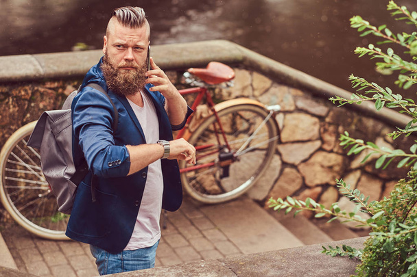 Portrait of a bearded male with a haircut dressed in casual clothes with a backpack, standing in a city park, talking by phone. - Foto, Bild