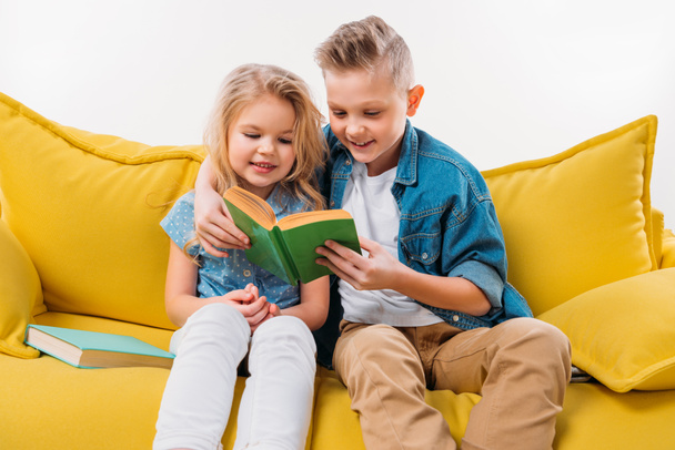 happy siblings reading book and sitting on yellow sofa - 写真・画像
