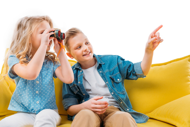 boy pointing while sister looking at binoculars, sitting on yellow sofa - Fotoğraf, Görsel