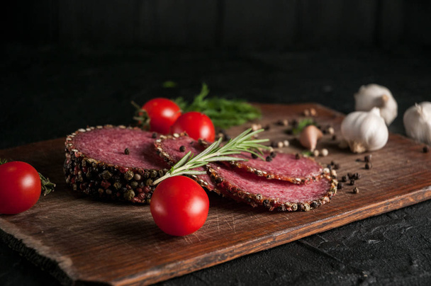 Smoked salami on a black table and a brown board. Sausages with rosemary, garlic and pepper and tomato cherry, dill, parsley. - Фото, изображение