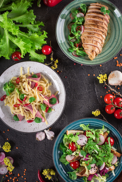 Delicious dinner. Italian pasta, fresh vegetable salad and roasted chicken breast on the black background. Healthy food. Close up. Top view - Photo, Image