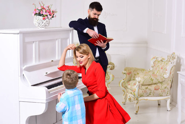 Child stand near piano keyboard, white interior background. Musician education concept. Mother teaches kid play piano, musical instrument. Boy adorable try to play piano, while parents watching him. - Photo, image