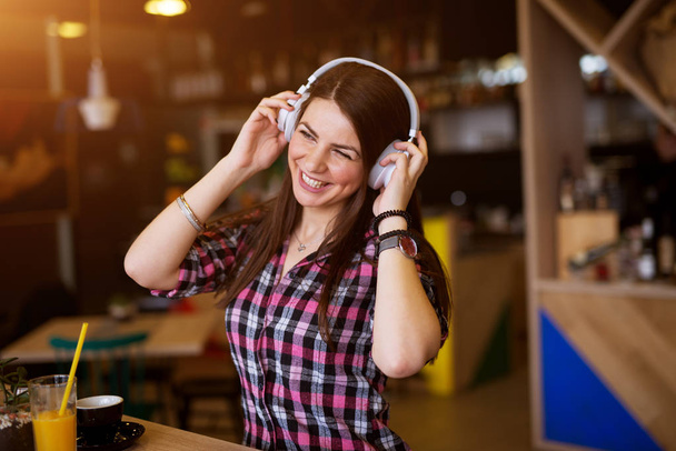 young cheerful woman enjoying music on headphones sitting at the table in cafe  - Fotó, kép