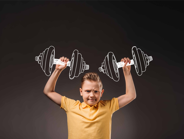 preteen boy pretending to be a sportsman and lifting big drawn dumbbells, isolated on grey - Фото, изображение