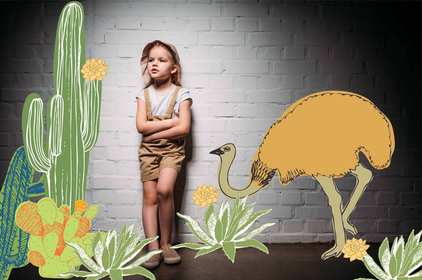 child in safari costume with crossed arms standing at white wall with cactuses and ostrich illustration - Photo, Image