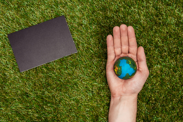 περικομμένη εικόνα του ανθρώπου που κρατώντας μοντέλο γη από πλευρά, μαυροπίνακα για την πράσινη χλόη, έννοια την ημέρα της γης - Φωτογραφία, εικόνα