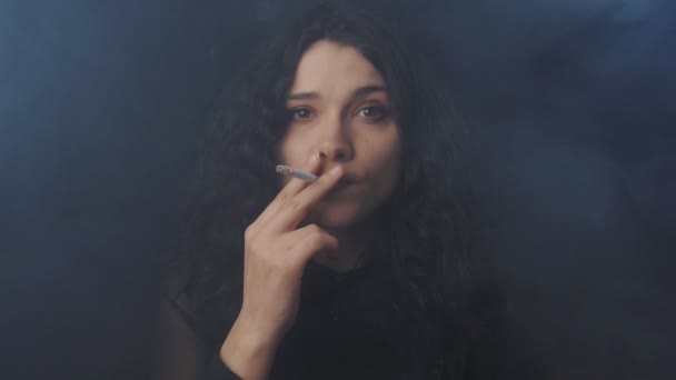 Portrét mladé ženy bruneta s kudrnatými vlasy kouřil cigaretu a při pohledu na fotoaparát v temné místnosti uzené - Záběry, video