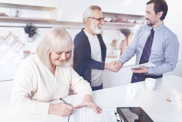 Well done. Cheeful delighted woman signing insurance contract while her husband shaking hands with agent in the background - Foto, Bild