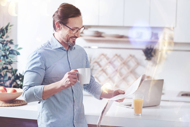 Daily press. Cheerful smiling adult man reading newspaper and drinking coffee while standing in the kitchen - Photo, Image