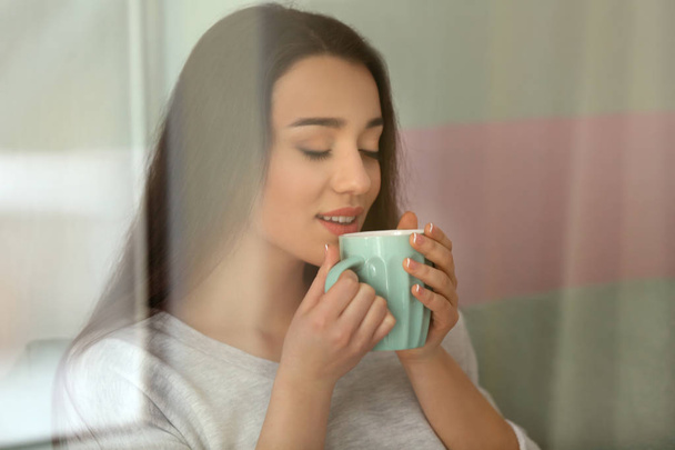 Όμορφη νεαρή γυναίκα με φλιτζάνι καυτό τσάι στο σπίτι, δείτε μέσα από το παράθυρο - Φωτογραφία, εικόνα