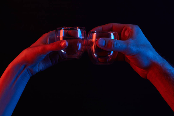 περικοπεί shot του άνδρα και της γυναίκας που τσούγκριζαν ποτήρια κάτω από ήπια φως που απομονώνονται σε μαύρο - Φωτογραφία, εικόνα