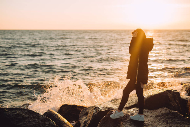 Mladá dívka stojí na kameny u moře na jaře. Zezadu je západ slunce a z toho to je svobodné a tiché - Fotografie, Obrázek