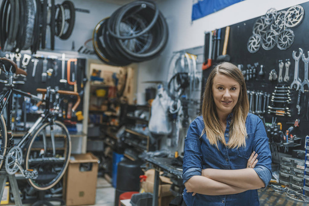 Portrait of female technician smiling and standing with folded arms in bicycle repair shop. - Фото, зображення
